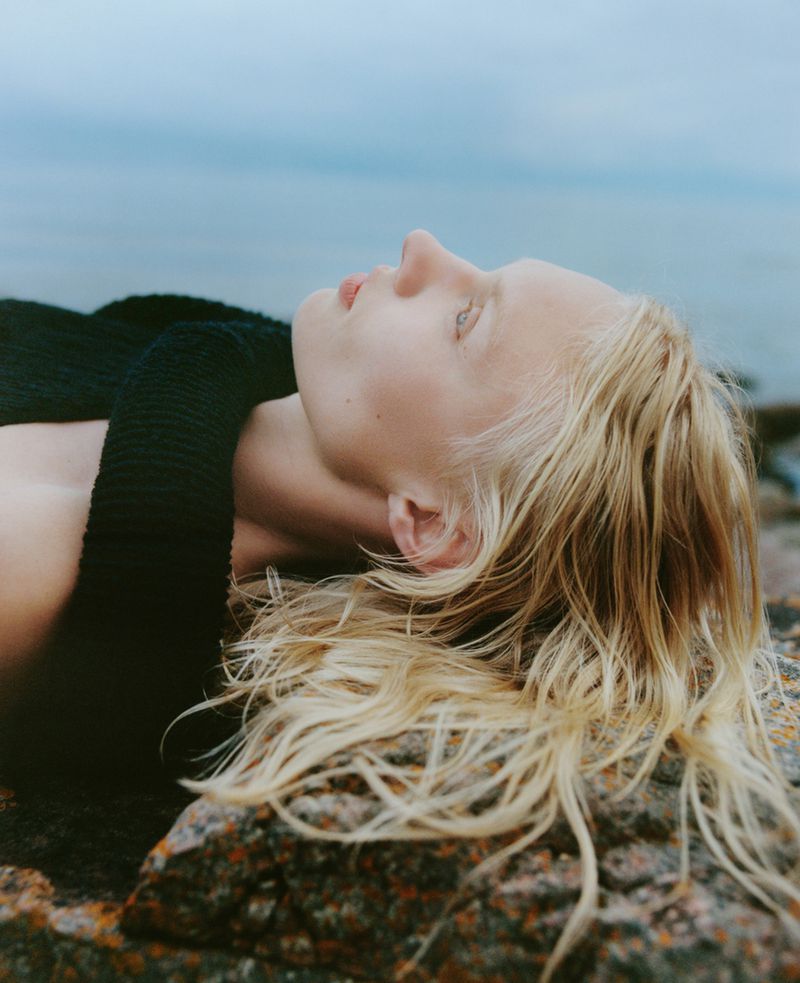 Eye-level shot of a woman lying on rocks near a body of water, her blonde hair spread out, wearing a black top.