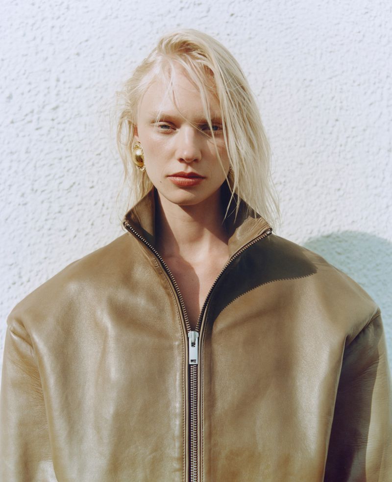Close-up of a model with blonde hair wearing a brown leather zip-up jacket against a white wall, showcasing fashion and style.