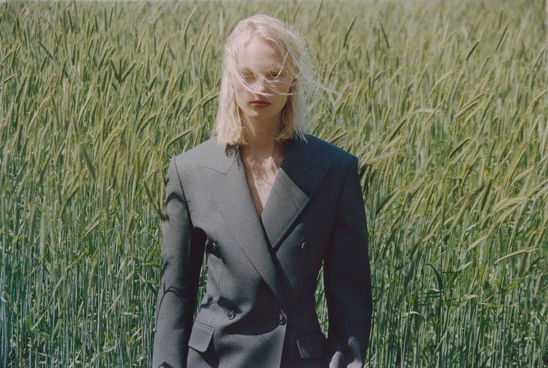Fashion shot of a blonde woman in a grey blazer standing in a field of wheat during daytime, highlighting natural beauty and fashion.