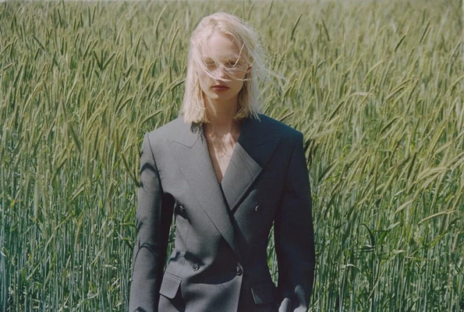 Fashion shot of a blonde woman in a grey blazer standing in a field of wheat during daytime, highlighting natural beauty and fashion.