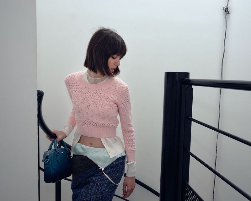 A woman in a pink sweater and blue skirt walks down stairs with a black handrail, holding a blue handbag in an indoor setting.