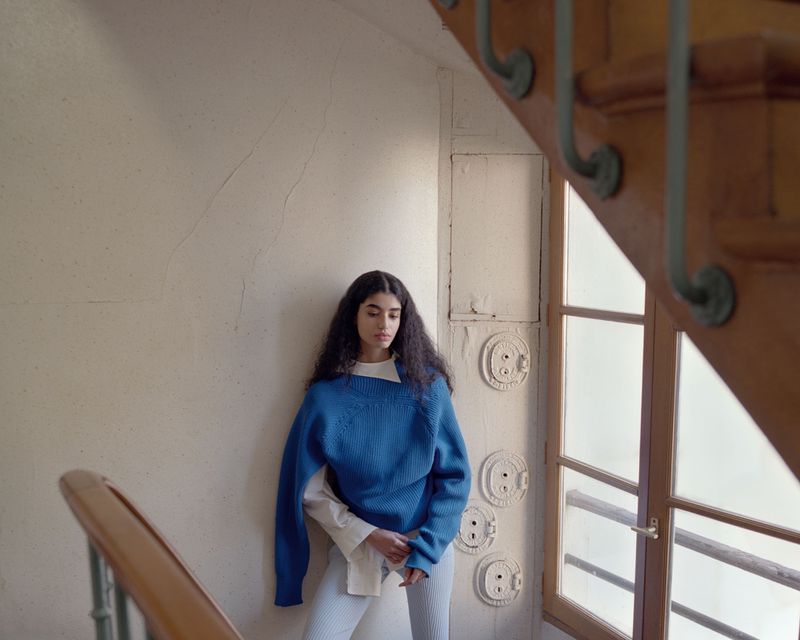 Indoor portrait of a young woman on a staircase, wearing a blue sweater, with natural light filtering through a nearby window.