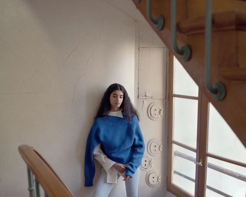Indoor portrait of a young woman on a staircase, wearing a blue sweater, with natural light filtering through a nearby window.