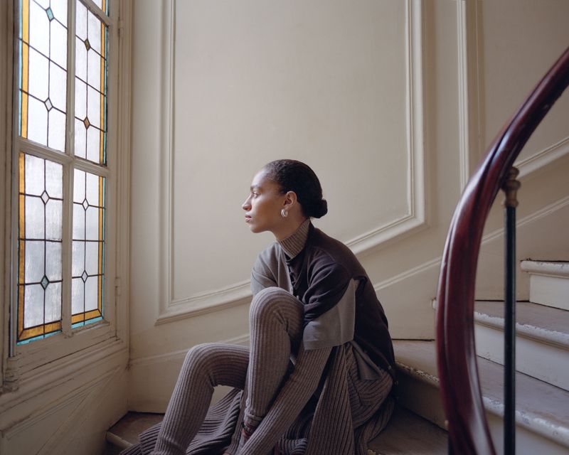 Young adult woman in brown knitwear sitting on an interior staircase looking out a stained glass window in a brightly lit interior.