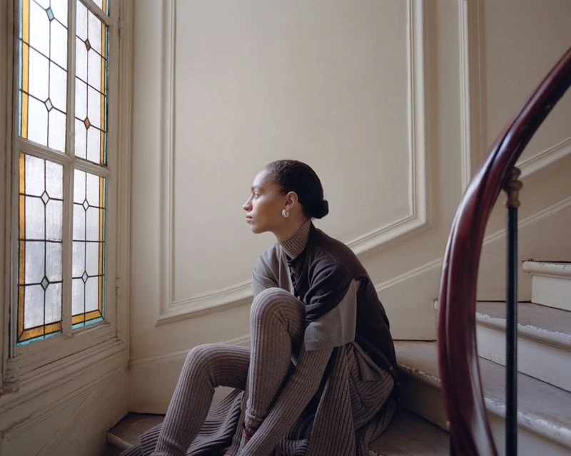 Young adult woman in brown knitwear sitting on an interior staircase looking out a stained glass window in a brightly lit interior.
