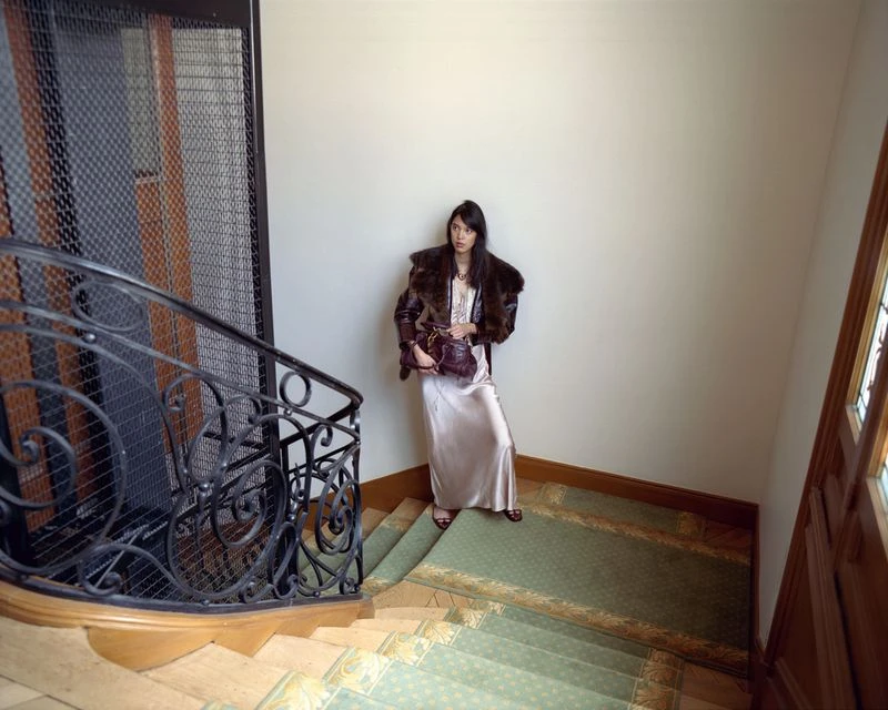 A woman with dark hair poses on a staircase with a vintage-style bag and coat. The scene is lit by natural light.