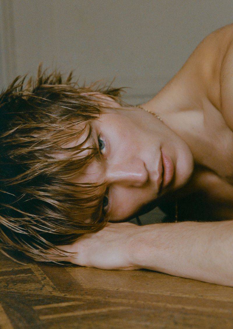 Portrait of a shirtless blonde man with light eyes laying on a wooden surface, looking at the camera in an indoor setting.