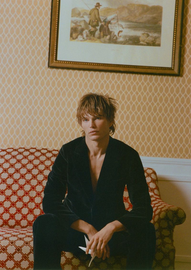 A young adult with blond hair sits on an ornate sofa in front of a patterned wallpaper, holding a paper origami bird in a medium shot.