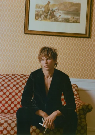 A young adult with blond hair sits on an ornate sofa in front of a patterned wallpaper, holding a paper origami bird in a medium shot.