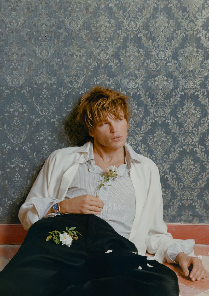 Portrait of a blonde male model in unbuttoned shirt with a flower, seated against a patterned wall with black pants on a tiled floor.