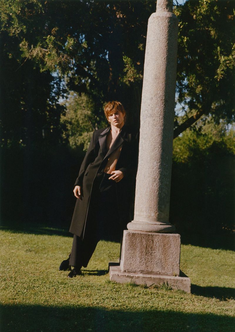 An adult male model poses in a black suit next to a stone pillar on a lawn with trees in the background, showcasing a fashion editorial style.