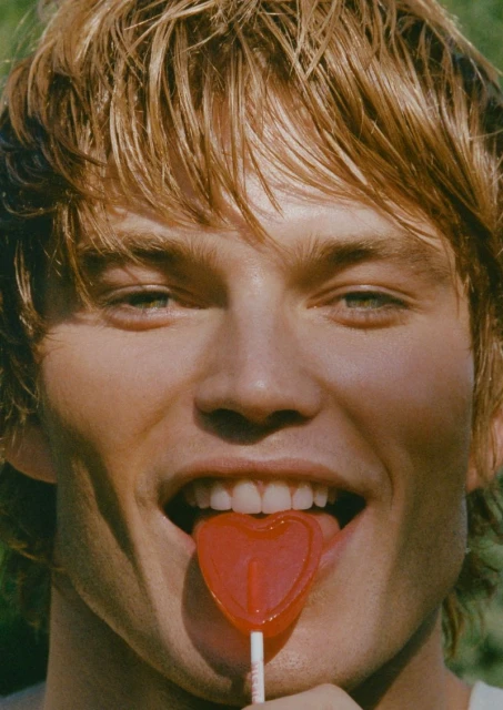 Close-up studio shot of a blonde adult male sticking out his tongue with a red heart-shaped lollipop against a green background.