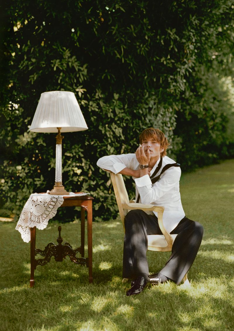 Blonde-haired person sitting in chair on grass near table lamp in outdoor garden setting with sunlight and shadows through trees.
