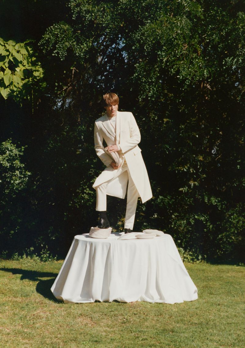 Man in a light-colored suit stands on a draped table outdoors, set against a backdrop of green foliage, captured in natural light.