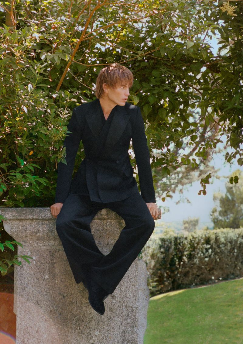 An adult male in a black suit poses outdoors, seated on a stone ledge with green foliage in the background, bathed in natural light.
