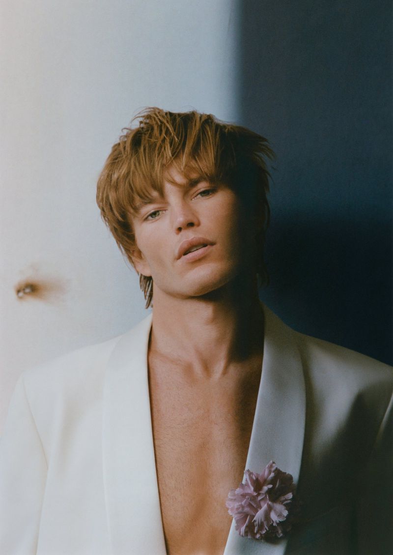 Close-up portrait of a fair-skinned adult male with reddish-blond hair wearing a white suit jacket with a flower pinned to the lapel.