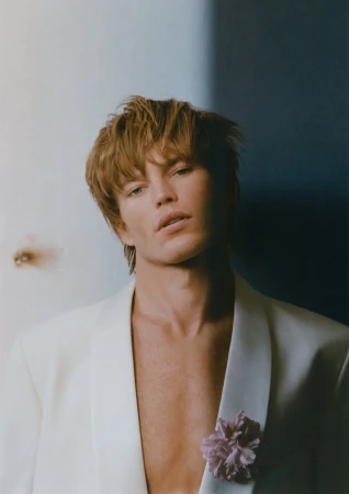 Close-up portrait of a fair-skinned adult male with reddish-blond hair wearing a white suit jacket with a flower pinned to the lapel.