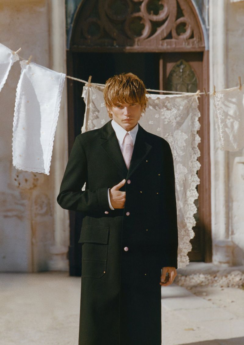 Adult male fashion model stands in an outdoor setting wearing a long black coat and pink tie with white linens hanging in the background.