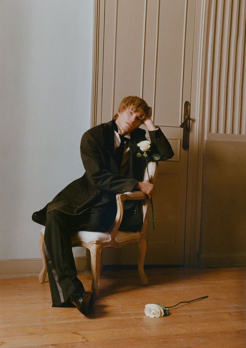 Young blond male fashion model poses in a tuxedo, seated on a chair with a white rose in hand in front of a paneled door.