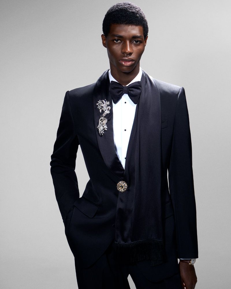 Portrait of a black male model in a black tuxedo with silver brooches and a bowtie against a light grey studio backdrop.