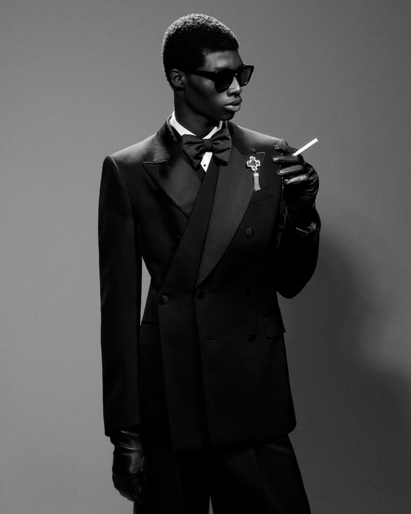 Black and white portrait of a dark-skinned adult male in a black suit, bow tie, and sunglasses, holding a cigarette in gloved hand.