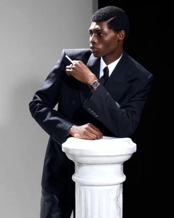 Portrait of a Black man in a black suit with cigarette, leaning on a marble pillar against a black and white backdrop.