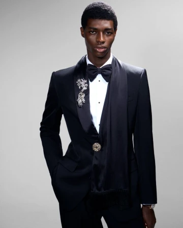 Black man wearing a black tuxedo with bow tie and brooch against a neutral grey studio background