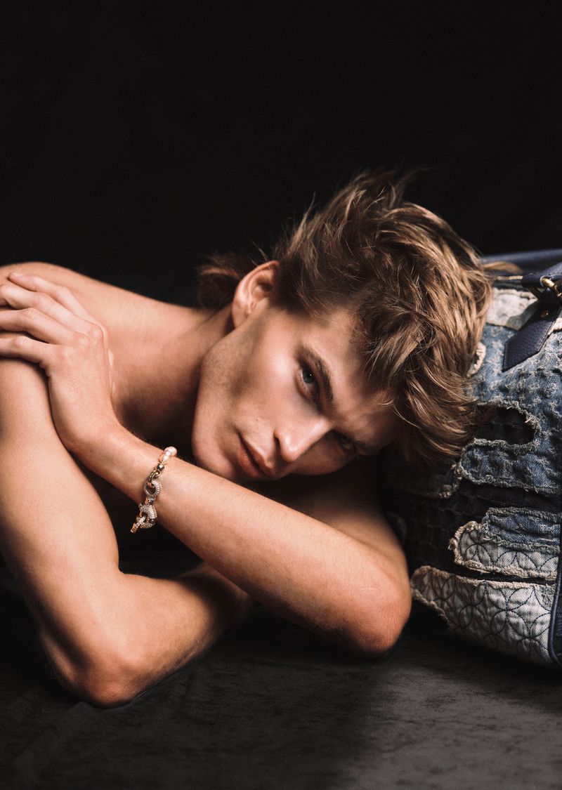 Handsome man poses shirtless with a slight smile next to a denim bag in a dimly lit studio with a dark gray surface.