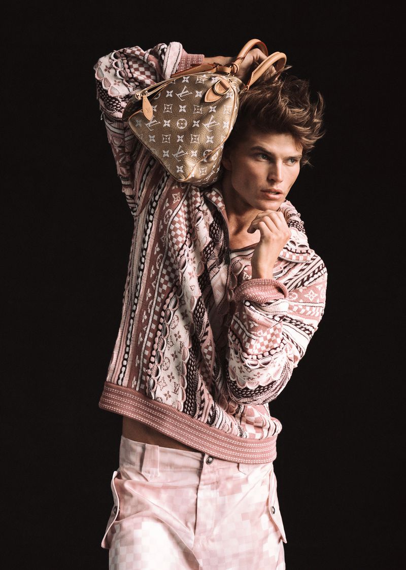 A tan skinned male fashion model with light brown hair poses with patterned shirt and pants. He is holding a Louis Vuitton bag.