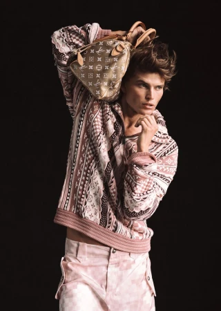 A tan skinned male fashion model with light brown hair poses with patterned shirt and pants. He is holding a Louis Vuitton bag.