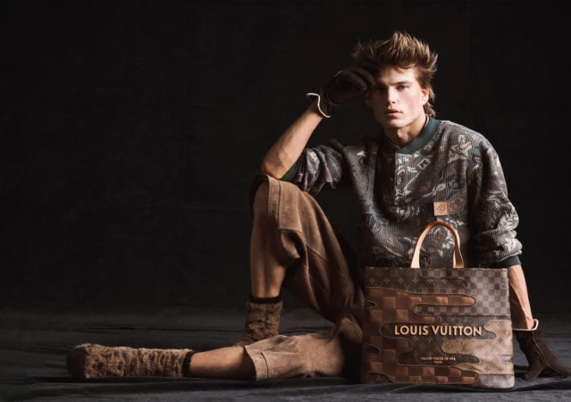 A young adult male fashion model poses while sitting on the floor with a Louis Vuitton handbag against a dark studio background.