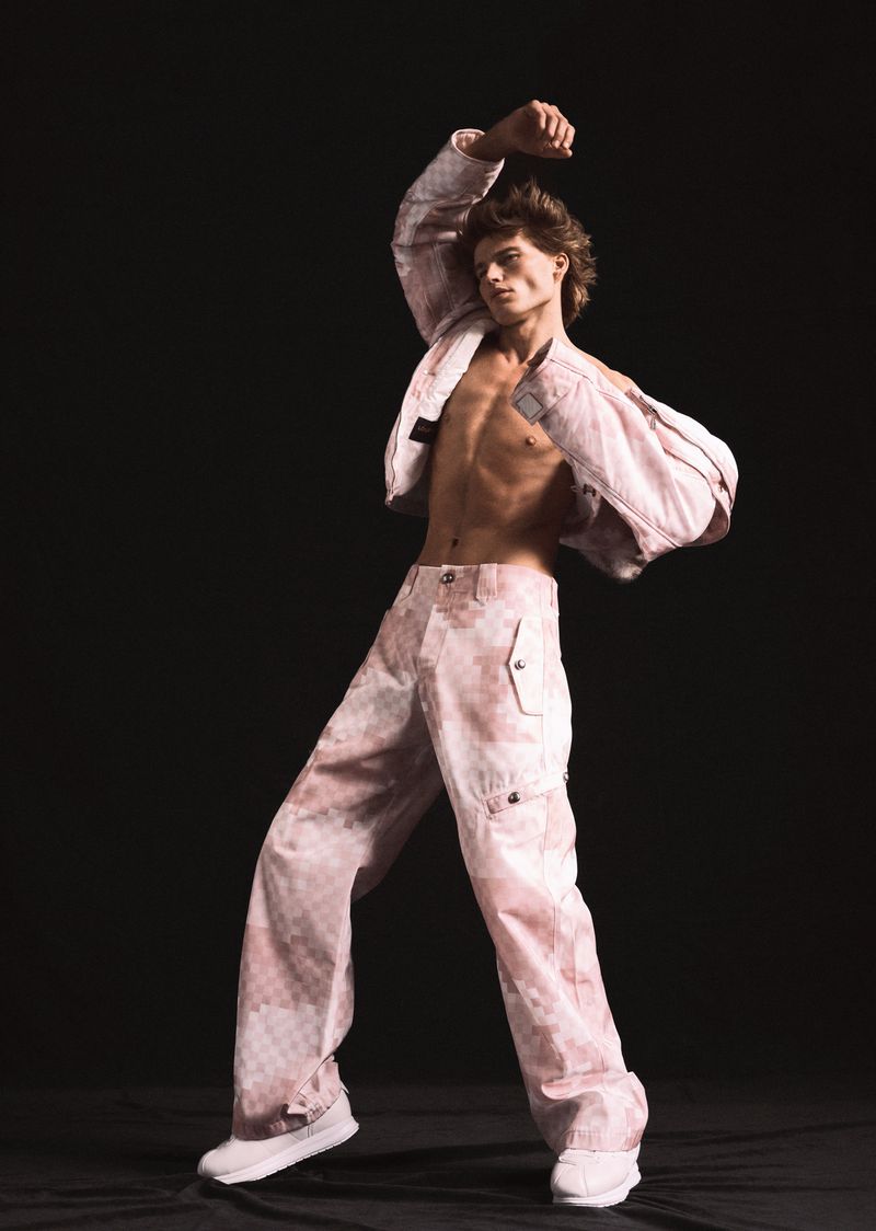Male model poses in a pink and white patterned jacket and baggy pants against a dark backdrop in a fashion photography studio.