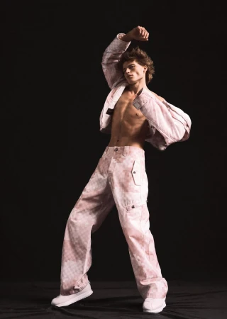 Male model poses in a pink and white patterned jacket and baggy pants against a dark backdrop in a fashion photography studio.