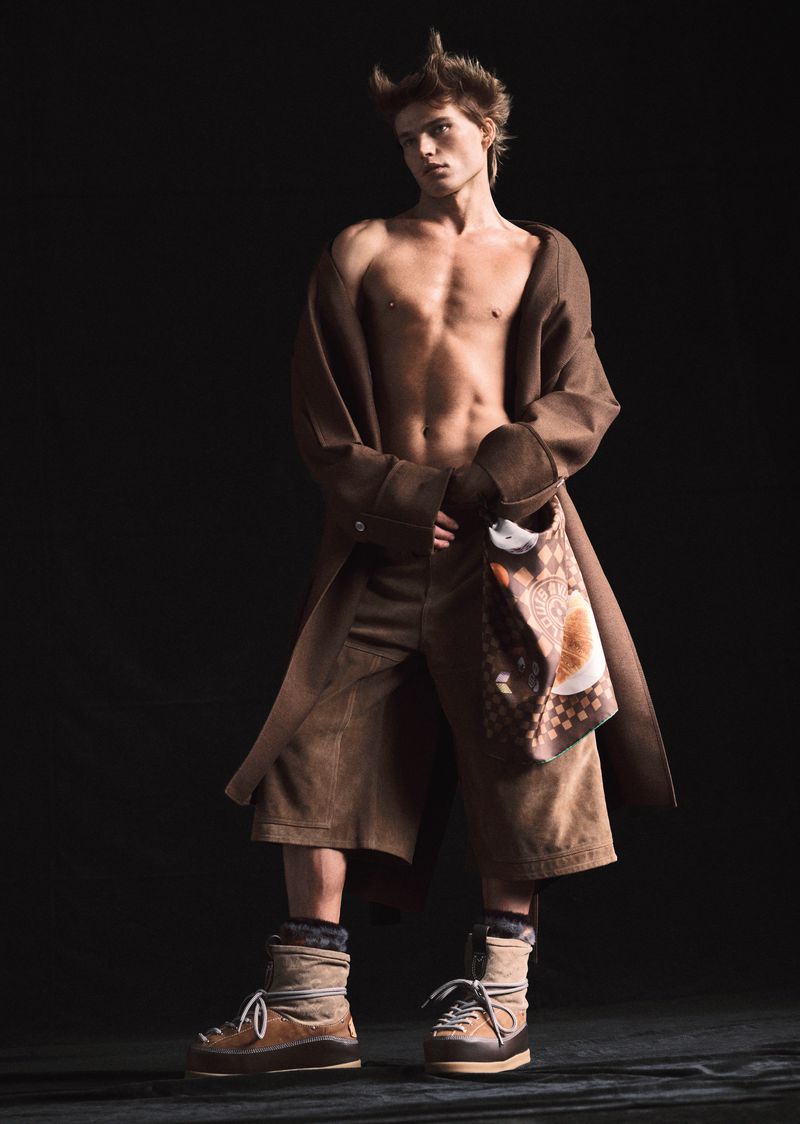 A fashion shot of a young adult male with bare chest, wearing brown shorts and a coat against a dark background in a photo studio.