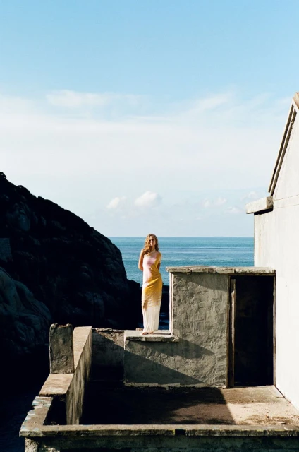 Woman standing on a concrete terrace overlooking the ocean on a sunny day, with a rocky coastline and blue sky in the background.
