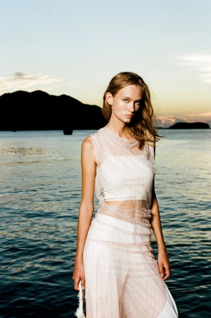 Fashion photo of a woman in a sheer top and pants standing by the ocean with a dark, silhouetted land mass in the background at sunset.