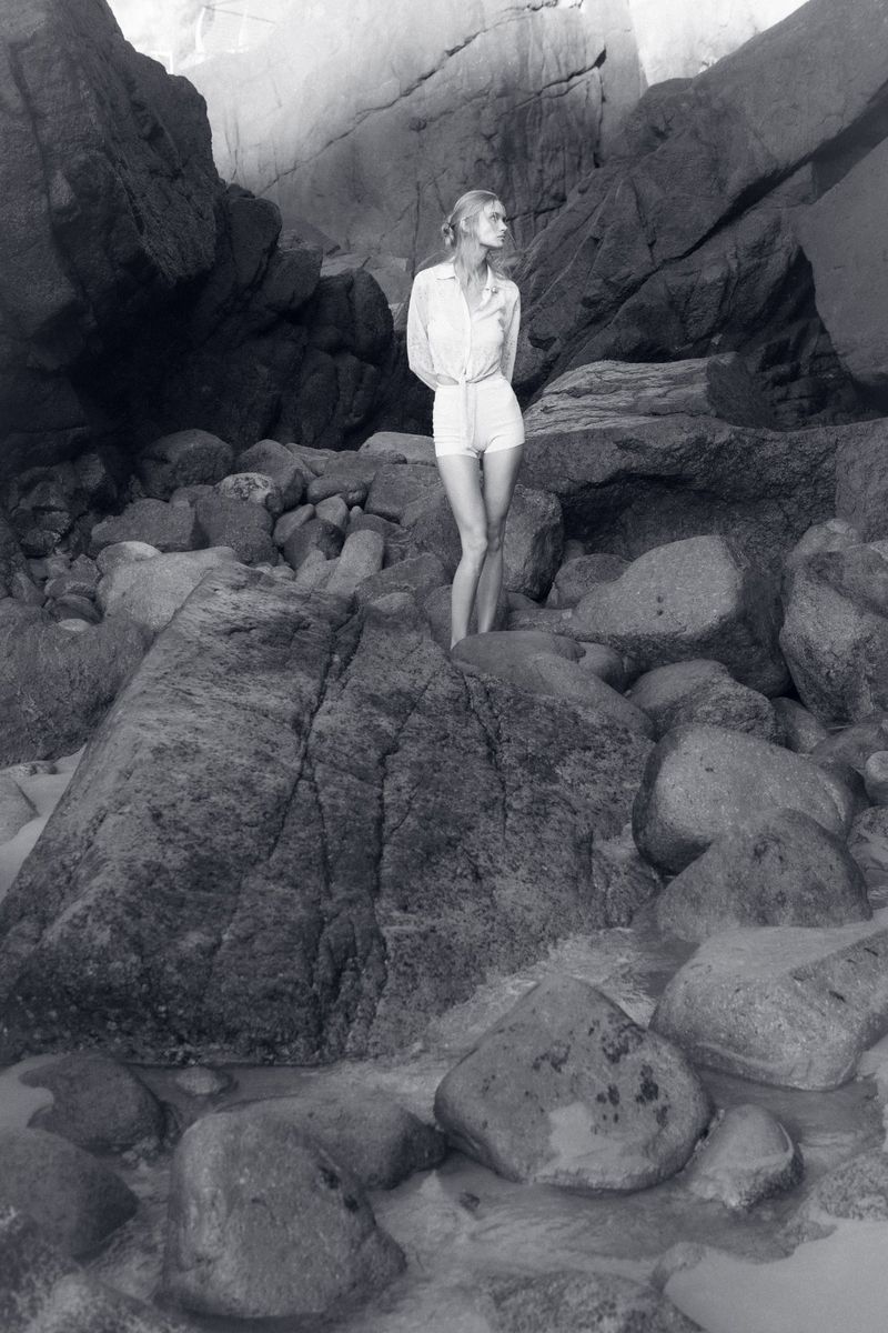 Model standing on a rocky beach wearing a light button-down shirt and shorts in a black and white editorial photograph.