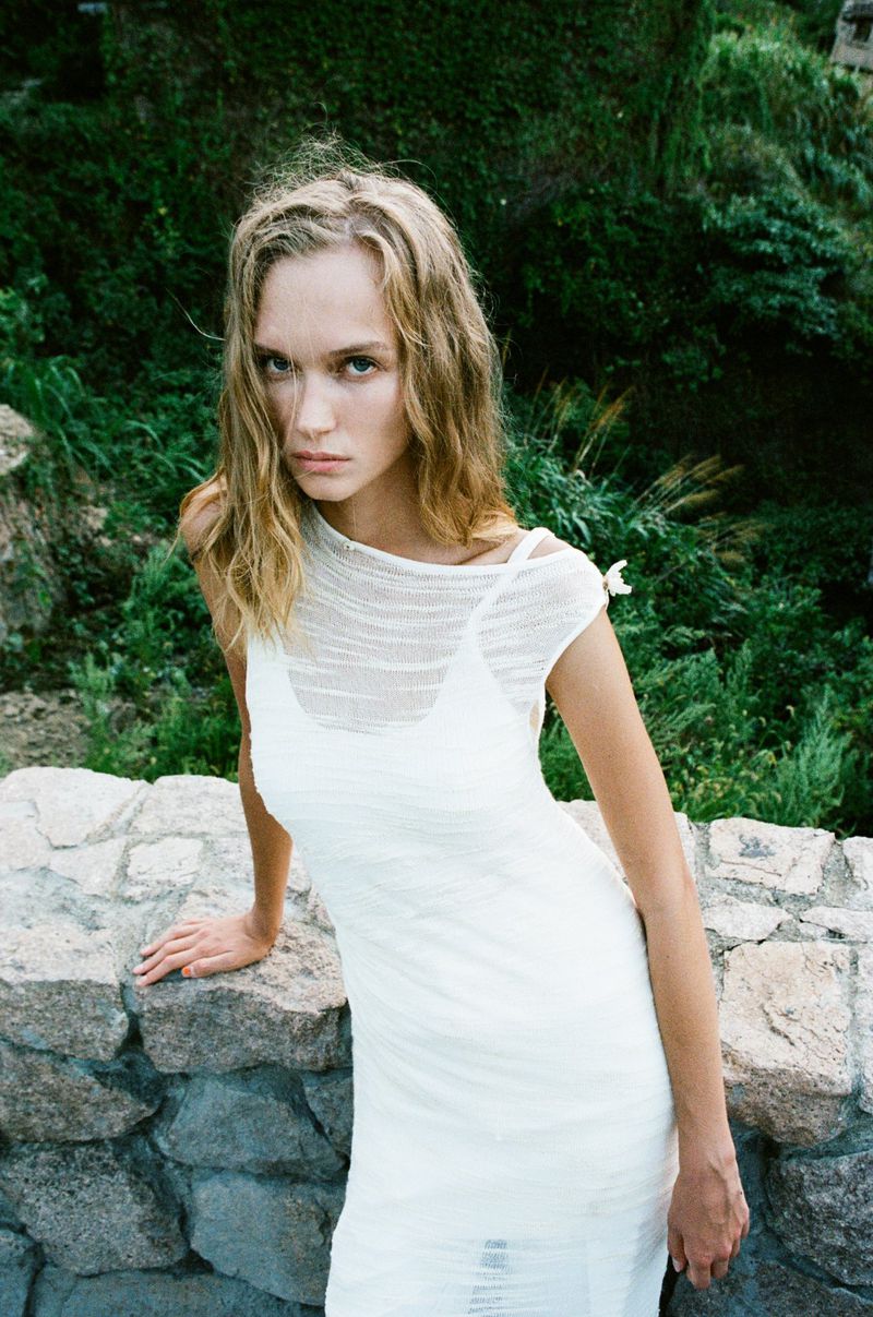 A slender woman with blonde hair poses outdoors wearing a white dress, set against a background of green foliage and a stone wall.