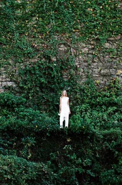 Woman in white clothing stands on an overgrown stone ledge surrounded by a wall covered in vines and greenery, forming a natural backdrop.