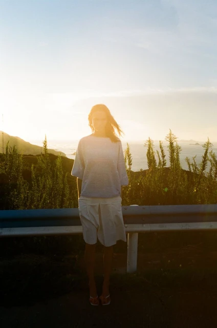 Woman in a white dress and shirt standing in front of a guardrail with a scenic view of the ocean and mountains at sunset.