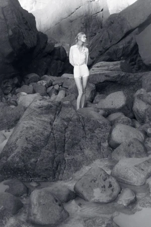 Model standing on a rocky beach wearing a light button-down shirt and shorts in a black and white editorial photograph.