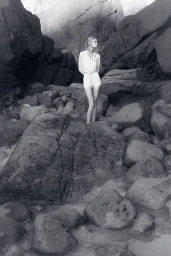 Model standing on a rocky beach wearing a light button-down shirt and shorts in a black and white editorial photograph.