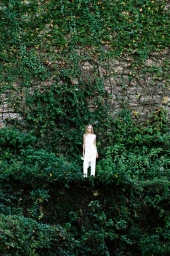 A model standing in a white dress against a massive stone wall covered in dense green ivy and foliage.