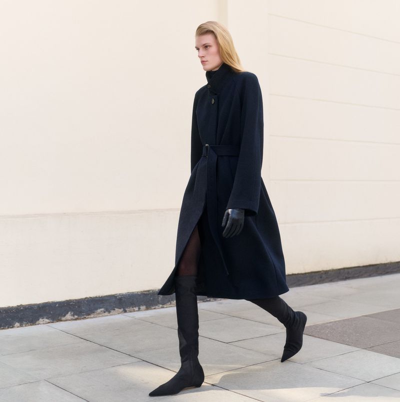 A full-length shot of a fair-skinned model in motion, wearing a long navy coat, gloves, and sock boots, against a light neutral outdoor background.