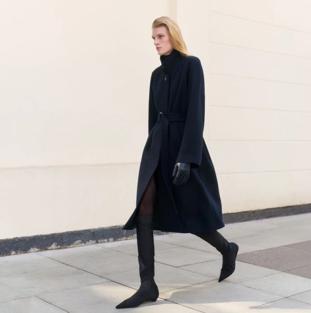 A full-length shot of a fair-skinned model in motion, wearing a long navy coat, gloves, and sock boots, against a light neutral outdoor background.