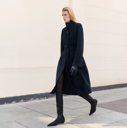 A full-length shot of a fair-skinned model in motion, wearing a long navy coat, gloves, and sock boots, against a light neutral outdoor background.