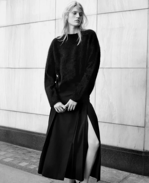 Monochrome fashion shot of a woman in a black sweater and slit skirt, posing against a textured wall in an outdoor setting.