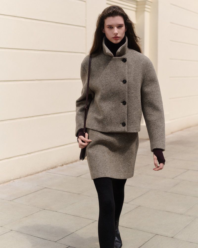 Model walking outdoors wearing a textured taupe wool jacket and skirt set with a dark shoulder bag