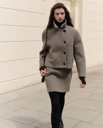Model walking outdoors wearing a textured taupe wool jacket and skirt set with a dark shoulder bag