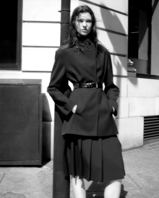 A black and white shot captures a stylish woman in a jacket and pleated skirt standing on a city street with textured walls.